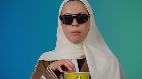 تعابير الارهاق والنعاس ، الاستمتاع بمشاهدة الأفلام داخل السينما، تصوير مقرب لامرأة عربية خليجية اماراتيه محجبة ترتدي عباءه وتحمل كيس فوشار والجوال ، التسلية والترفيه بقضاء أوقات ممتعة، ايماءات السعادة والسرور لمتابعة الفيلم المفضل، خلفية ملونه