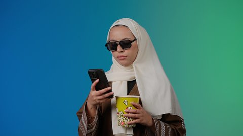 التسلية والترفيه بقضاء أوقات ممتعة، متابعة الفيلم المفضل، تعابير الارهاق والنعاس ، الاستمتاع بمشاهدة الأفلام داخل السينما، تصوير مقرب لامرأة عربية خليجية اماراتيه محجبة ترتدي عباءه وتحمل كيس فوشار والجوال ، خلفية ملونه