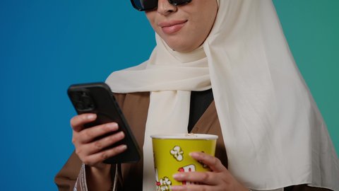 الاستمتاع بمشاهدة الأفلام داخل السينما، تصوير مقرب لامرأة عربية خليجية اماراتيه محجبة ترتدي عباءه وتحمل كيس فوشار والجوال ، التسلية والترفيه بقضاء أوقات ممتعة، ايماءات السعادة والسرور لمتابعة الفيلم المفضل، خلفية ملونه