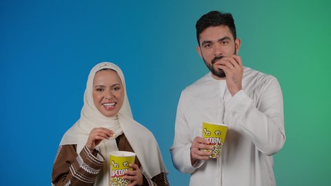تناول الفوشار والمشروبات الباردة اثناء متابعة الأفلام والمسلسلات، قضاء وقت ممتع في السينما، تصوير مقرب لزوجيين عربيين خليجيين اماراتيين مبتسميين ينظران الى الكاميرا بتعابير السرور والسعادة، الاستمتاع بالأنشطة العائلية الترفيهيه، خلفية ملونه