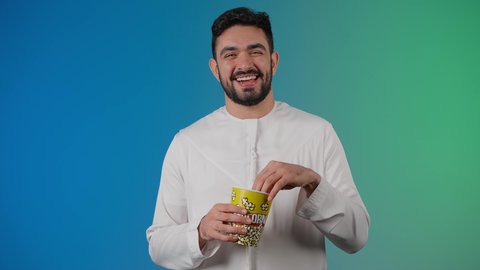 مفهوم التسليه والترفيه، تعابير المرح والاستمتاع ، قضاء أوقات ممتعة ومسلية، تصوير مقرب لرجل عربي خليجي اماراتي يرتدي الزى التقليدي يحمل بيده علبة الفشار ويقوم بمشاهده الافلام ، مشاهدة فيلم سينمائي ، خلفيه ملونه
