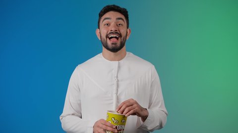 تعابير المرح والاستمتاع ، قضاء أوقات ممتعة ومسلية، مفهوم التسليه والترفيه، تصوير مقرب لرجل عربي خليجي اماراتي يرتدي الزى التقليدي يحمل بيده علبة الفشار ويقوم بمشاهده الافلام ، مشاهدة فيلم سينمائي ، خلفيه ملونه