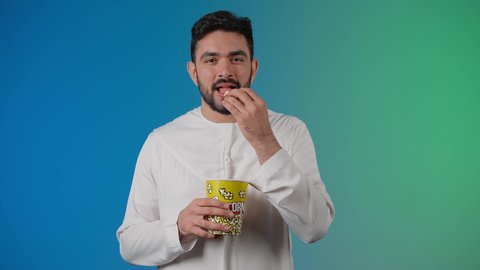 رفع اصبع السبابه بايماءات الموافقه ، تعابير الجودة والامتياز والرضي ، قضاء أوقات ممتعة ومسلية، مفهوم التسليه والترفيه، تصوير مقرب لرجل عربي خليجي اماراتي يرتدي الزى التقليدي يحمل بيده علبة الفشار ويقوم بمشاهده الافلام ، مشاهدة فيلم سينمائي ، خلفيه ملونه