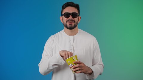 رفع قبضه اليد بايماءات الفرح والحماس ، قضاء أوقات ممتعة ومسلية، رجل عربي خليجي اماراتي يرتدي الكندوره والغتره يحمل بيده علبة الفشار ومستمتع بمشاهدة الأفلام ، إيماءات وجه ويدين تدل على السعادة والمتعة، مشاهدة فيلم سينمائي، مفهوم التسليه والترفيه