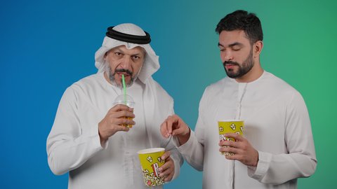 Having a good time eating popcorn while watching TV, the concept of group entertainment, watching movies and plays in the cinema, a picture of two Emirati Gulf Arab friends wearing kandora and ghutra watching movies, holding cups full of popcorn in their hands, expressions of laughter and joking, drinking cold drinks