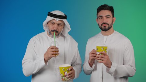 تعابير الضحك والهزار ، مفهوم التسليه والترفيه الجماعيه ، مشاهدة الأفلام والعروض المسرحية في السينما، تصوير لصديقان عربيان خليجيان امارتيان يرتديان الكندوره والغتره يشاهدان الافلام ، يحملان بيدهما أكواب مليئه بالبوب كورن، شرب المشروبات البارده، قضاء وقت ممتع بتناول الفشار اثناء مشاهده التلفاز