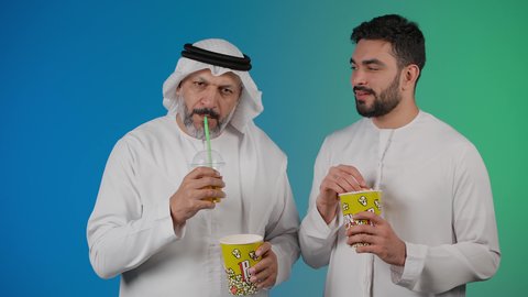 The concept of group entertainment and recreation, drinking cold drinks, watching movies and plays in the cinema, spending a pleasant time eating popcorn while watching TV, a picture of two Emirati Gulf Arab friends wearing kanduras and ghutras watching movies, holding cups full of popcorn in their hands