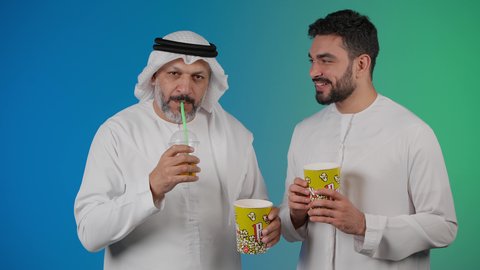 شرب المشروبات البارده ، مشاهدة الأفلام والعروض المسرحية في السينما، قضاء وقت ممتع بتناول الفشار اثناء مشاهده التلفاز ، تصوير لصديقان عربيان خليجيان امارتيان يرتديان الكندوره والغتره يشاهدان الافلام ، يحملان بيدهما أكواب مليئه بالبوب كورن، مفهوم التسليه والترفيه الجماعيه