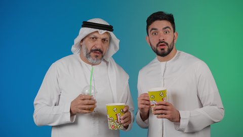 تعابير الصدمه والذهول، مشاهدة الأفلام والعروض المسرحية في السينما، قضاء وقت ممتع بتناول الفشار اثناء مشاهده التلفاز ، تصوير لصديقان عربيان خليجيان امارتيان يرتديان الكندوره والغتره يشاهدان الافلام ، يحملان بيدهما أكواب مليئه بالبوب كورن، مفهوم التسليه والترفيه الجماعيه