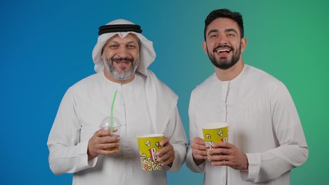 مشاهدة الأفلام والعروض المسرحية في السينما، تعابير السعاده والسرور، قضاء وقت ممتع بتناول الفشار اثناء مشاهده التلفاز ، تصوير لصديقان عربيان خليجيان امارتيان يرتديان الكندوره والغتره يشاهدان الافلام ، يحملان بيدهما أكواب مليئه بالبوب كورن، مفهوم التسليه والترفيه الجماعيه
