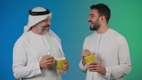 تعابير السعاده والسرور، قضاء وقت ممتع بتناول الفشار اثناء مشاهده التلفاز، مفهوم التسليه والترفيه الجماعيه ، تصوير لصديقان عربيان خليجيان امارتيان يرتديان الكندوره والغتره يشاهدان الافلام ، يحملان بيدهما أكواب مليئه بالبوب كورن