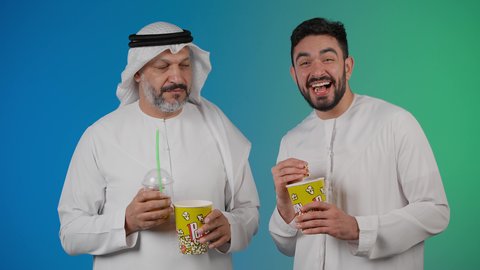 Having a good time eating popcorn while watching TV, the concept of group entertainment and fun, a picture of two Emirati Gulf Arab friends wearing kandura and ghutra watching movies, holding cups full of popcorn in their hands, expressions of happiness and pleasure