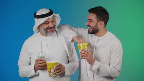 مفهوم التسليه والترفيه الجماعي, قضاء وقت ممتع بتناول الفشار اثناء مشاهده التلفاز، تصوير لصديقان عربيان خليجيان امارتيان يرتديان الكندوره والغتره يشاهدان الافلام ، يحملان بيدهما أكواب مليئه بالبوب كورن، تعابير  السعاده والسرور