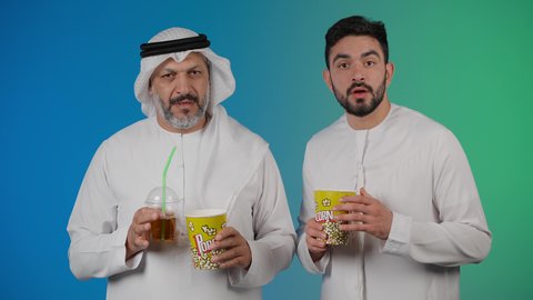 مشاهدة الأفلام والعروض المسرحية في السينما، قضاء وقت ممتع بتناول الفشار اثناء مشاهده التلفاز ، تصوير لصديقان عربيان خليجيان امارتيان يرتديان الكندوره والغتره يشاهدان الافلام بتعابير المفاجأه ، يحملان بيدهما أكواب مليئه بالبوب كورن، مفهوم التسليه والترفيه الجماعيه
