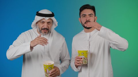 محاوله رؤيه شئ بعيد ، مشاهدة الأفلام والعروض المسرحية في السينما، قضاء وقت ممتع بتناول الفشار اثناء مشاهده التلفاز ، تصوير لصديقان عربيان خليجيان امارتيان يرتديان الكندوره والغتره يشاهدان الافلام ، يحملان بيدهما أكواب مليئه بالبوب كورن، مفهوم التسليه والترفيه الجماعيه