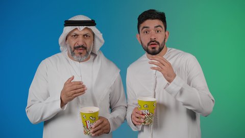 ايماءات الذهول والمفاجأه ، مشاهدة الأفلام والعروض المسرحية في السينما، قضاء وقت ممتع بتناول الفشار اثناء مشاهده التلفاز ، تصوير لصديقان عربيان خليجيان امارتيان يرتديان الكندوره والغتره يشاهدان الافلام ، يحملان بيدهما أكواب مليئه بالبوب كورن، مفهوم التسليه والترفيه الجماعيه