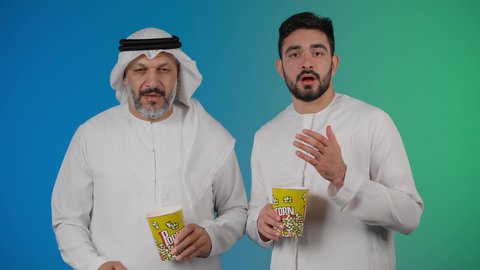 ايماءات النعاس والارهاق ، مشاهدة الأفلام والعروض المسرحية في السينما، قضاء وقت ممتع بتناول الفشار اثناء مشاهده التلفاز ، تصوير لصديقان عربيان خليجيان امارتيان يرتديان الكندوره والغتره يشاهدان الافلام ، يحملان بيدهما أكواب مليئه بالبوب كورن، مفهوم التسليه والترفيه الجماعيه