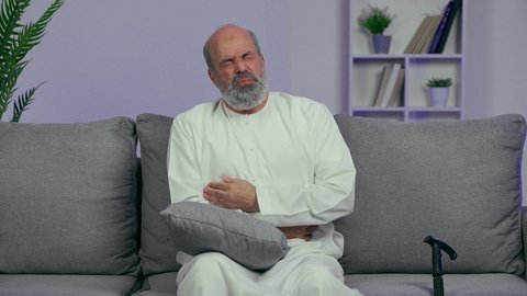 الانطواء على النفس من شدة الألم والتعب، ضرورة الراحة وأخذ العلاج المناسب، الشعور بالمرض ووجع في البطن، رجل كبير السن عربي خليجي إماراتي يرتدي الكندورة يضع يديه على بطنه بتعابير التألم، أوجاع القولون