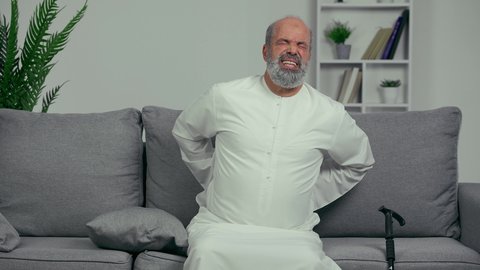 رجل عربي اماراتي كبير بالسن يضع يده على ظهره يشعر بحالة من الوجع والألم, شايب اماراتي يرتدي الكندورة, مسن يشعر بالتعب والألم الشديد في ظهره, مفهوم تعابيرالوجه ولغة الجسد