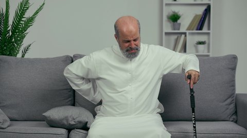 رجل عربي اماراتي كبير بالسن يضع يده على ظهره يشعر بحالة من الوجع والألم, شايب اماراتي يرتدي الكندورة يجلس ويمسك بالعصا, مسن يشعر بالتعب والألم الشديد في ظهره, مفهوم تعابيرالوجه ولغة الجسد