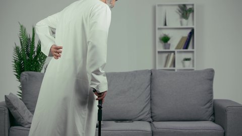 آلام الظهر والعمود الفقري، رجل عربي اماراتي كبير بالسن يضع يده على ظهره يشعر بحالة من الوجع والألم, شايب اماراتي يرتدي الكندورة يجلس ويمسك بالعصا, مسن يشعر بالتعب والألم الشديد في ظهره, مفهوم تعابيرالوجه ولغة الجسد