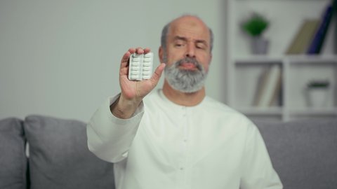 رجل كبير بالسن عربي خليجي اماراتي يرتدي الكندوره يجلس علي اريكه ويحمل شريط الدواء، ايماءه وجه منزعجه، مريض عربي اماراتي خليجي مسن يقوم بتلقي العلاج في المستشفى, الاهتمام بالصحة والالتزام بالأوقات الصحيحة لأخذ العلاج, استخدام عقار الدواء بوصفة طبية