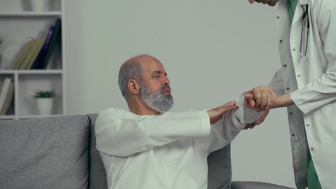 مريض عربي اماراتي كبير بالسن يشكو من آلام في الكتف واليد,  طبيب اماراتي يرتدي المعطف الابيض يقوم بفحص المريض والتأكد من سلامة اليد, صحة وأطباء, حالة مرضية, مهن ووظائف طبيه، ايماءه التعب الشديد