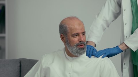 الحاجة للعلاج الفيزيائي، الإحساس بالتعب والإرهاق, وجع مفاصل و عظام الرقبة, آلام العنق والعمود الفقري, طبيب عربي خليجي إماراتي يرتدي المعطف الابيض والقفزات يقوم بفحص رجل مسن، رجل مسن يرتدي كندوره يعاني من آلام الفقرات