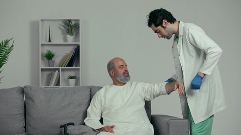 طبيب اماراتي يرتدي القفازات والمعطف الطبي يقوم بأخذ عينة دم من رجل مسن، تقديم الرعاية الصحية والخدمات، ايماءه وجه تدل علي الارهاق، التعقيم والاهتمام بصحه المريض، رجل مسن اماراتي يرتدي كندوره بيضاء يجلس علي اريكه رماديه يقوم بميد يداه لسحب عينه دم