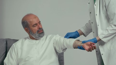 طبيب اماراتي يرتدي القفازات والمعطف الطبي يقوم بأخذ عينة دم من رجل مسن، تقديم الرعاية الصحية والخدمات، ايماءه وجه تدل علي الارهاق، التعقيم والاهتمام بصحه المريض، رجل مسن اماراتي يرتدي كندوره بيضاء يجلس علي اريكه رماديه يقوم بميد يداه لسحب عينه دم