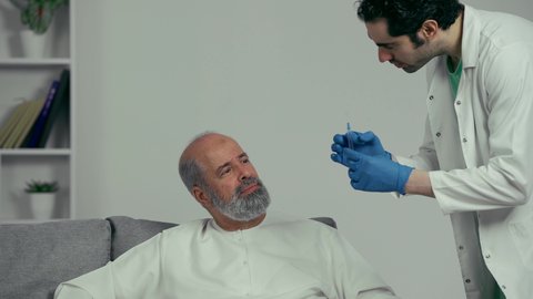 طبيب اماراتي يرتدي القفازات والمعطف الطبي يقوم باعطاء ابرة لرجل كبير بالسن عربي خليجي اماراتي يرتدي الكندوره، تقديم الرعاية الصحية والخدمات، ايماءه وجه تدل علي الارهاق الشديد
