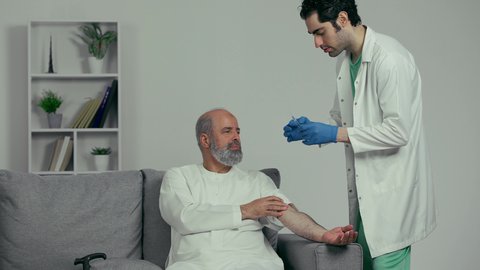 طبيب اماراتي يرتدي القفازات والمعطف الطبي يقوم باعطاء ابرة لرجل كبير بالسن عربي خليجي اماراتي يرتدي الكندوره، تقديم الرعاية الصحية والخدمات، ايماءه وجه تدل علي الارهاق الشديد