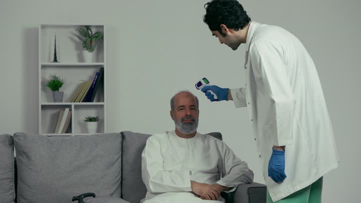 Working in the medical sector, an Emirati doctor holding a digital infrared thermometer examines an elderly patient, following preventive and safety measures against the Corona virus, health awareness and avoiding the spread of infection.