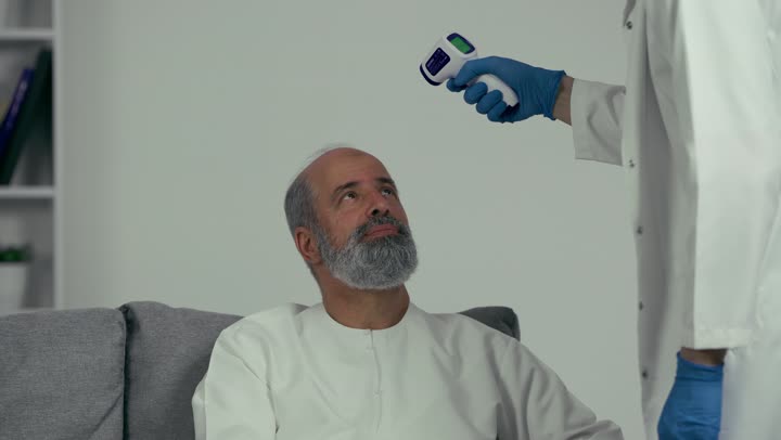Working in the medical sector, an Emirati doctor holding a digital infrared thermometer examines an elderly patient, following preventive and safety measures against the Corona virus, health awareness and avoiding the spread of infection.