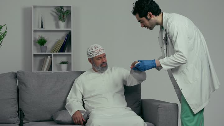 The concept of medicine and healthcare, an Arab Gulf Emirati doctor wearing a white coat and gloves is measuring the blood pressure using an electronic scale for an elderly patient wearing a white cap and kandura, using modern medical equipment.