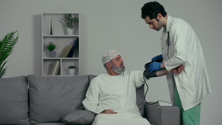Using modern medical equipment, the concept of medicine and healthcare, an Arab Gulf Emirati doctor wearing a white coat and gloves measuring the blood pressure of an elderly patient wearing a white cap and kandura