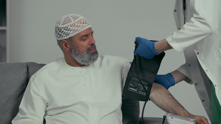 Using modern medical equipment, the concept of medicine and healthcare, an Arab Gulf Emirati doctor wearing a white coat and gloves measuring the blood pressure of an elderly patient wearing a white cap and kandura