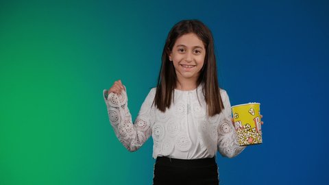 القفز عاليا بتعابير الحماس والفرح، مفهوم الترفيه عن النفس والتسلية ، ايماءات الاستمتاع بمشاهدة فيلم ممتع، متابعة الأفلام والعروض المسرحية في السينما، الابتعاد عن روتين الحياة اليومية، تصوير لبنت عربية خليجية اماراتيه مبتسمة ترتدي الزي الكاجوال ونظاره سوداء تمسك بيدها علبه الفوشار ، خلفية ملونه