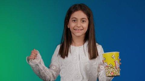 ايماءات الاستمتاع بمشاهدة فيلم ممتع، مفهوم الترفيه عن النفس والتسلية ،القفزعاليا بتعابير الحماس والفرح، متابعة الأفلام والعروض المسرحية في السينما، الابتعاد عن روتين الحياة اليومية، تصوير لبنت عربية خليجية اماراتيه مبتسمة ترتدي الزي الكاجوال ونظاره سوداء تمسك بيدها علبه الفوشار ، خلفية ملونه