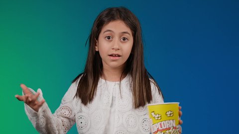 Expressions of astonishment and surprise, interest in elegance and outward appearance, getting away from the routine of daily life, gestures of enjoying watching an enjoyable movie, following movies and theatrical performances in the cinema, a picture of a smiling Emirati Gulf Arab girl wearing casual clothes and black glasses holding a box of popcorn in her hand, a colorful background