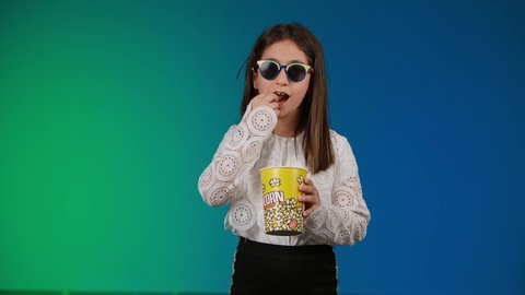الابتعاد عن روتين الحياة اليومية، ايماءات الاستمتاع بمشاهدة فيلم ممتع، متابعة الأفلام والعروض المسرحية في السينما، تصوير لبنت عربية خليجية اماراتيه مبتسمة ترتدي الزي الكاجوال ونظاره سوداء تمسك بيدها علبه الفوشار ، خلفية ملونه
