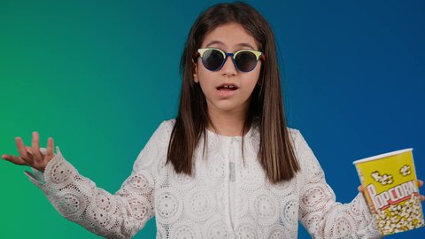 جهل امر ما ، تعابير عدم المعرفه والحيره ،متابعة الأفلام والعروض المسرحية في السينما، الابتعاد عن روتين الحياة اليومية،  ايماءات الاستمتاع بمشاهدة فيلم ممتع، تصوير لبنت عربية خليجية اماراتيه مبتسمة ترتدي الزي الكاجوال ونظاره سوداء تمسك بيدها علبه الفوشار ، خلفية ملونه