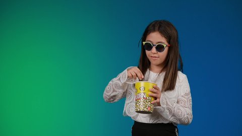 الابتعاد عن روتين الحياة اليومية،  متابعة الأفلام والعروض المسرحية في السينما، ايماءات الاستمتاع بمشاهدة فيلم ممتع، تصوير لبنت عربية خليجية اماراتيه مبتسمة ترتدي الزي الكاجوال ونظاره سوداء تمسك بيدها علبه الفوشار ، خلفية ملونه