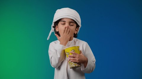 الإحساس بالإرهاق و الحاجه الى النوم والراحه ,تصوير لولد عربي اماراتي خليجي يرتدي الكندورة والغتره يقف بإيماءات تدل على النعاس يمسك بيده علبه الفوشار, متابعة الأفلام والعروض المسرحية في السينما، الابتعاد عن روتين الحياة اليومية، مفهوم الترفيه عن النفس والتسلية