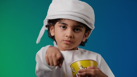 الاشاره الى شئ ما ، متابعة الأفلام والعروض المسرحية في السينما، مفهوم الترفيه عن النفس والتسلية ,تصوير لولد عربي اماراتي خليجي يرتدي الكندورة والغتره يمسك بيده علبه الفوشار,  الابتعاد عن روتين الحياة اليومية