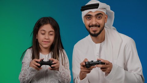 ألعاب ترفيهية، اللعب الجماعي, قضاء وقت ممتع مع العائله, إيماءات وجه تدل على الفوز والخساره، المتعة و التسلية بألعاب الفيديو، اب وابنته عربيان اماراتيان خليجيان يمسكان عصا التحكم و يلعبان بألعاب الفيديو، تسليه وترفيه, تقنيات و ألعاب
