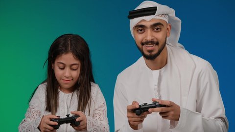 إيماءات وجه تدل على الفوز والخساره، قضاء وقت ممتع مع العائله,  المتعة و التسلية بألعاب الفيديو، اب وابنته عربيان اماراتيان خليجيان يمسكان عصا التحكم و يلعبان بألعاب الفيديو، ألعاب البلايستيشن الترفيهية، اللعب الجماعي, تسليه وترفيه, تقنيات و ألعاب