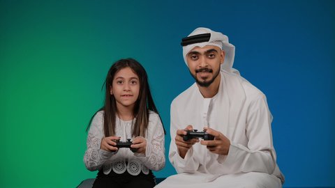 تقنيات و ألعاب، اللعب الجماعي, قضاء وقت ممتع مع العائله, إيماءات وجه تدل على الفوز والخساره، المتعة و التسلية بألعاب الفيديو، اب وابنته عربيان اماراتيان خليجيان يمسكان عصا التحكم و يلعبان بألعاب الفيديو، تسليه وترفيه