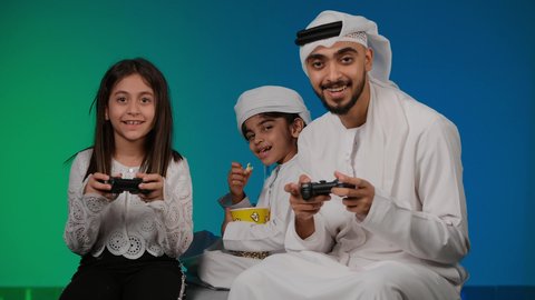 ايماءات وحركات يدين تدل على التحدي باللعبة، مفهوم الانشطة الترفيهية المسلية مع الأهل، تصوير لعائلة عربية خليجية اماراتيه تقضي أوقات ممتعة في لعب الألعاب الالكترونية، أجواء عائلية مليئة بالسعادة والحب، خلفيه ملونه