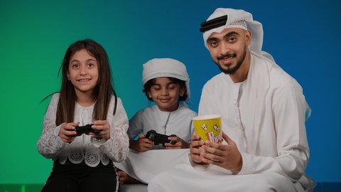 أجواء عائلية مليئة بالسعادة والحب، ايماءات وحركات يدين تدل على التحدي باللعبة، مفهوم الانشطة الترفيهية المسلية مع الأهل، تصوير لعائلة عربية خليجية اماراتيه تقضي أوقات ممتعة في لعب الألعاب الالكترونية، خلفيه ملونه